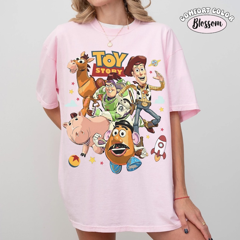 May include: A light pink Comfort Colors t-shirt featuring a colorful graphic of Toy Story characters, including Woody, Buzz Lightyear, and Mr. Potato Head. The words "TOY STORY" are printed above the characters. The shirt has short sleeves.