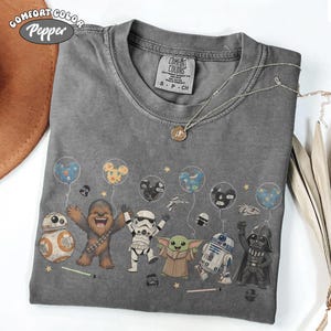 May include: Gray Comfort Colors t-shirt featuring Star Wars characters with balloons. The design includes BB-8, Chewbacca, a Stormtrooper, Grogu, R2-D2, and Darth Vader. The shirt has a crew neck and short sleeves.