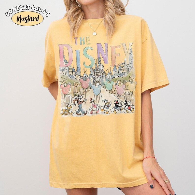 Pu&ograve; includere: T-shirt color senape con la scritta "THE DISNEY" in lettere pastello. La grafica presenta personaggi Disney che attraversano una strada con un castello sullo sfondo e palloncini di Topolino.
