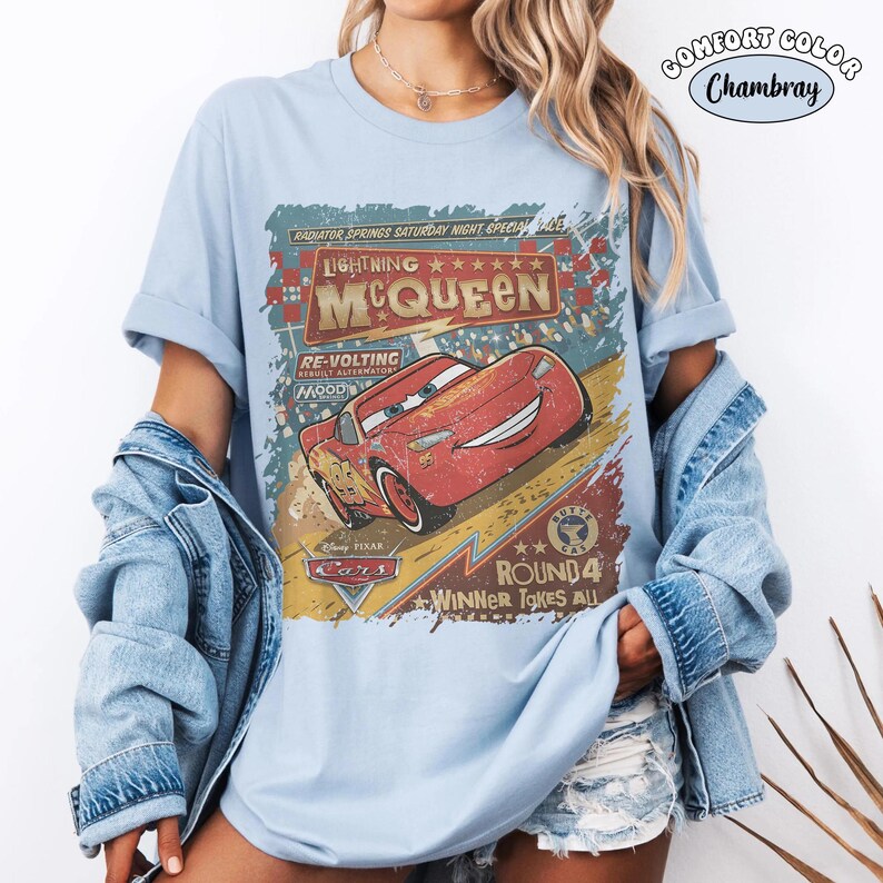 May include: Light blue t-shirt featuring a distressed graphic of Lightning McQueen from the Disney Pixar Cars movie. The design includes text that reads "Lightning McQueen" and "Winner Takes All". The shirt is labeled "Comfort Color Chambray".