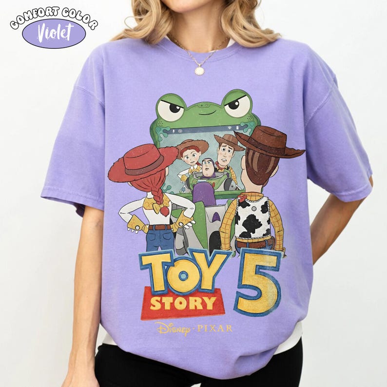 Pu&ograve; includere: Una maglietta Comfort Colors viola con una grafica del film Disney Pixar Toy Story 5. Il design include i personaggi Woody, Buzz Lightyear, Jessie e altri. La maglietta ha un girocollo e maniche corte.