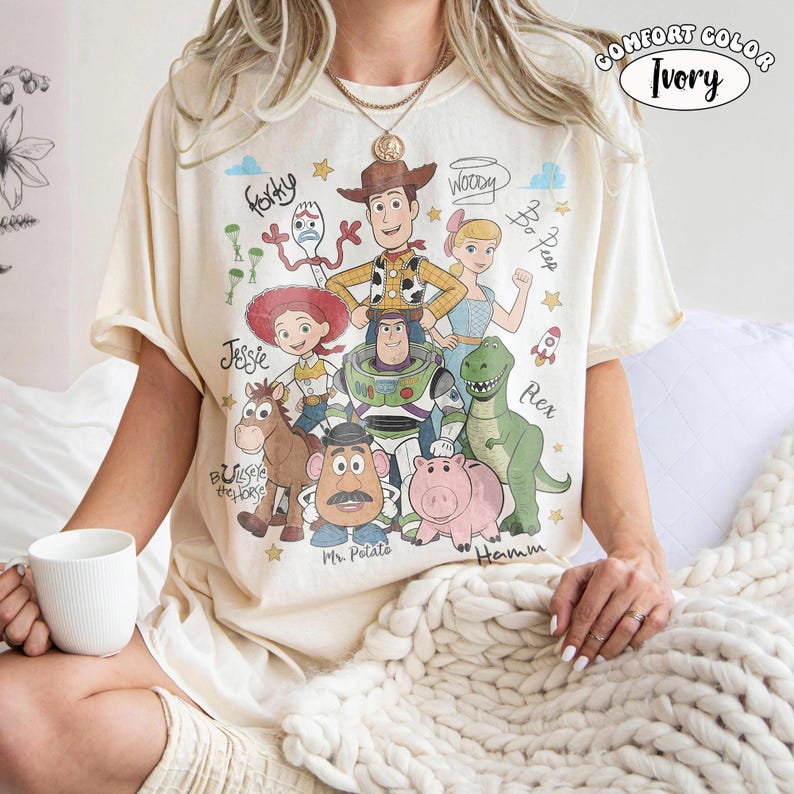 Może przedstawiać: Koszulka w kolorze kości słoniowej z nadrukiem graficznym przedstawiającym postacie z Toy Story, w tym Chudego, Buzza Astrala, Jessie, Pastereczkę, Rexa, Hamm, Celka, Pana Bulwę i Sztućka, z ich imionami zapisanymi kursywą i małymi gwiazdkami.