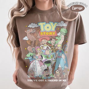 K&ouml;nnte beinhalten: Ein espresso-braunes T-Shirt mit einem Grafikdruck von Toy Story-Figuren wie Woody, Buzz Lightyear und Jessie. Der Text TOY STORY steht oben und YOU'VE GOT A FRIEND IN ME unten. Ein Comfort Color-Etikett ist auf dem T-Shirt zu sehen.