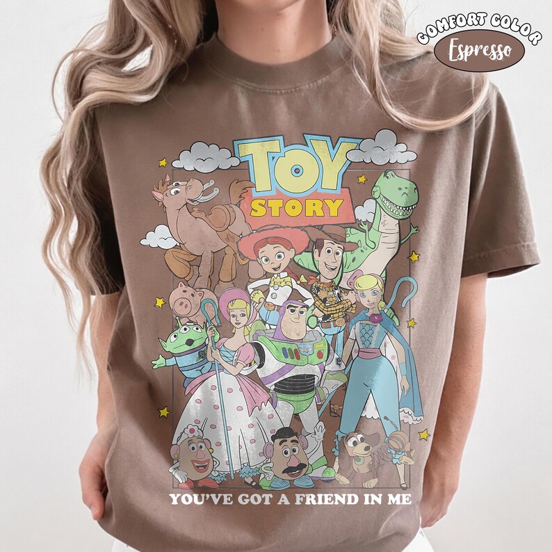K&ouml;nnte beinhalten: Ein espresso-braunes T-Shirt mit einem Grafikdruck von Toy Story-Figuren wie Woody, Buzz Lightyear und Jessie. Der Text TOY STORY steht oben und YOU'VE GOT A FRIEND IN ME unten. Ein Comfort Color-Etikett ist auf dem T-Shirt zu sehen.
