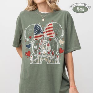 May include: Olive green t-shirt featuring a graphic of a castle with an American flag bow and heart accents. The shirt has the words "COMFORT COLOR Moss" printed on the upper right side. The design is in white, red, and blue.