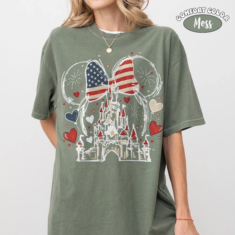 May include: Olive green t-shirt featuring a graphic of a castle with an American flag bow and heart accents. The shirt has the words "COMFORT COLOR Moss" printed on the upper right side. The design is in white, red, and blue.