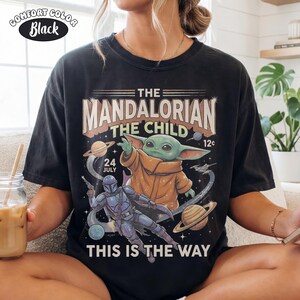 May include: A black short-sleeve t-shirt featuring a graphic print of The Mandalorian and The Child in space. The design includes planets, stars, and text that reads THE MANDALORIAN, THE CHILD, 24 JULY, 12c, and THIS IS THE WAY. A small label reads COMFORT COLOR Black.