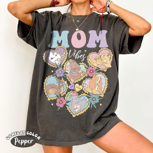 Pu&ograve; includere: T-shirt Comfort Colors grigio scuro con le parole "MOM Vibes" in colori pastello. La maglietta presenta illustrazioni a forma di cuore di personaggi Disney, circondate da fiori. La parola "Pepper" &egrave; stampata in basso sulla maglietta.