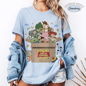 K&ouml;nnte beinhalten: Hellblaues T-Shirt mit einer Grafik von Charakteren aus Toy Story. Das Design zeigt eine Box mit dem Text "YOU'VE GOT A FRIEND IN ME SINCE 1995". Das Shirt hat hochgekrempelte &Auml;rmel und ist aus Comfort Colors Chambray.