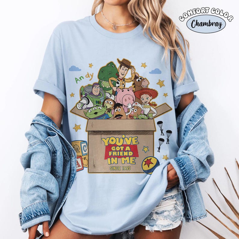 K&ouml;nnte beinhalten: Hellblaues T-Shirt mit einer Grafik von Charakteren aus Toy Story. Das Design zeigt eine Box mit dem Text "YOU'VE GOT A FRIEND IN ME SINCE 1995". Das Shirt hat hochgekrempelte &Auml;rmel und ist aus Comfort Colors Chambray.