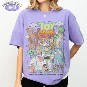 K&ouml;nnte beinhalten: Ein violettes Kurzarm-T-Shirt mit einem Grafikdruck der Toy Story-Figuren, darunter Woody, Buzz Lightyear, Jessie und Rex. Der Text TOY STORY und YOU'VE GOT A FRIEND IN ME ist im Design enthalten. Ein Comfort Color Violet-Etikett ist oben links zu sehen.