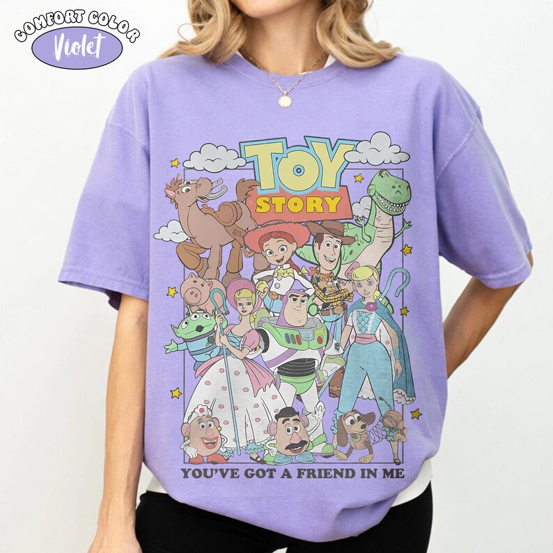 K&ouml;nnte beinhalten: Ein violettes Kurzarm-T-Shirt mit einem Grafikdruck der Toy Story-Figuren, darunter Woody, Buzz Lightyear, Jessie und Rex. Der Text TOY STORY und YOU'VE GOT A FRIEND IN ME ist im Design enthalten. Ein Comfort Color Violet-Etikett ist oben links zu sehen.