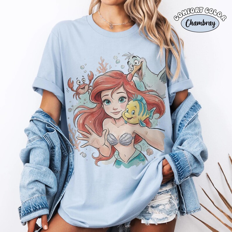 May include: Pale blue Comfort Colors Chambray t-shirt with a watercolour graphic of Ariel, Flounder, Sebastian, and Scuttle. The shirt has rolled-up sleeves and is worn with a denim jacket. The graphic is from The Little Mermaid.