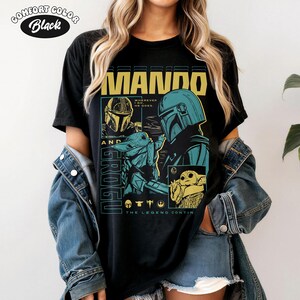 K&ouml;nnte beinhalten: Schwarzes T-Shirt mit einem Grafikdesign mit dem Text "MANDO AND GROGU". Das Design enth&auml;lt Illustrationen von Charakteren aus der Star Wars-Serie in Gelb-, T&uuml;rkis- und Blaut&ouml;nen. Der Text "THE LEGEND CONTINUES" ist ebenfalls vorhanden.