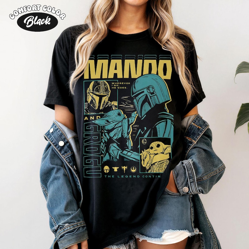 K&ouml;nnte beinhalten: Schwarzes T-Shirt mit einem Grafikdesign mit dem Text "MANDO AND GROGU". Das Design enth&auml;lt Illustrationen von Charakteren aus der Star Wars-Serie in Gelb-, T&uuml;rkis- und Blaut&ouml;nen. Der Text "THE LEGEND CONTINUES" ist ebenfalls vorhanden.