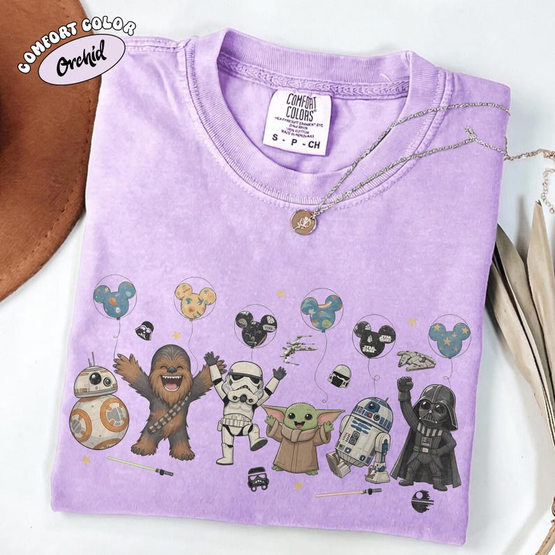 May include: A light purple Comfort Colors t-shirt featuring Star Wars characters and Mickey Mouse balloons. The shirt includes BB-8, Chewbacca, a Stormtrooper, Grogu, R2-D2, and Darth Vader. A silver necklace with a pendant is also visible.