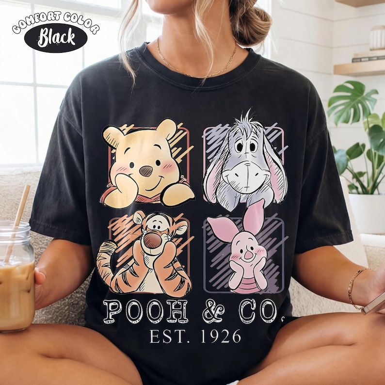 May include: Black t-shirt featuring four cartoon character portraits: Winnie the Pooh, Eeyore, Tigger, and Piglet. The shirt has the text "POOH & CO. EST. 1926" and the words "Comfort Color Black" in a circle.