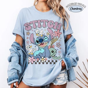 Puede incluir: Camiseta azul claro con la palabra "STITCH" en una fuente retro, con un personaje de dibujos animados, flores y patos. La camiseta tiene las mangas enrolladas y una etiqueta "Comfort Color Chambray".