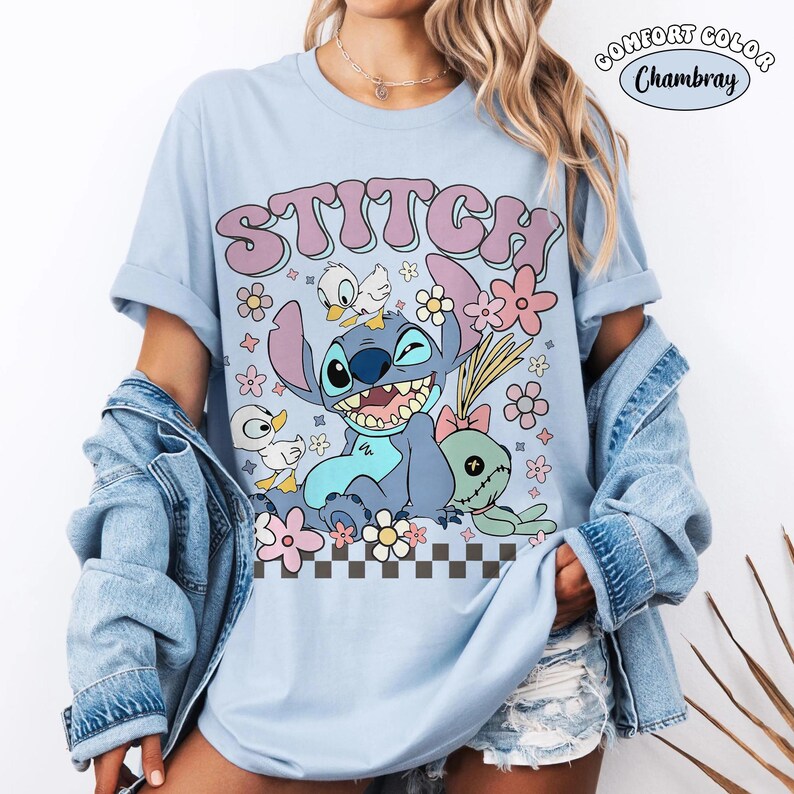 Puede incluir: Camiseta azul claro con la palabra "STITCH" en una fuente retro, con un personaje de dibujos animados, flores y patos. La camiseta tiene las mangas enrolladas y una etiqueta "Comfort Color Chambray".