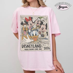 Puede incluir: Una camiseta rosa claro con un estampado gr&aacute;fico rectangular de personajes de Disney que incluye a Mickey Mouse, Minnie Mouse, el Pato Donald, Pluto y Goofy. El texto dice Disneyland Where Dreams Come True 2026, con firmas de personajes.