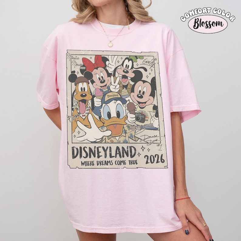 Puede incluir: Una camiseta rosa claro con un estampado gr&aacute;fico rectangular de personajes de Disney que incluye a Mickey Mouse, Minnie Mouse, el Pato Donald, Pluto y Goofy. El texto dice Disneyland Where Dreams Come True 2026, con firmas de personajes.