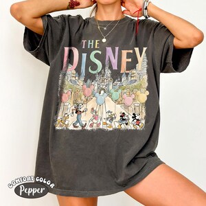 Pu&ograve; includere: T-shirt grigio scuro con grafica colorata. Il testo "THE DISNEY" &egrave; sopra un'illustrazione di personaggi Disney e un castello. La maglietta ha un aspetto vintage e sbiadito, con le parole "COMFORT COLOR Pepper" stampate in basso.