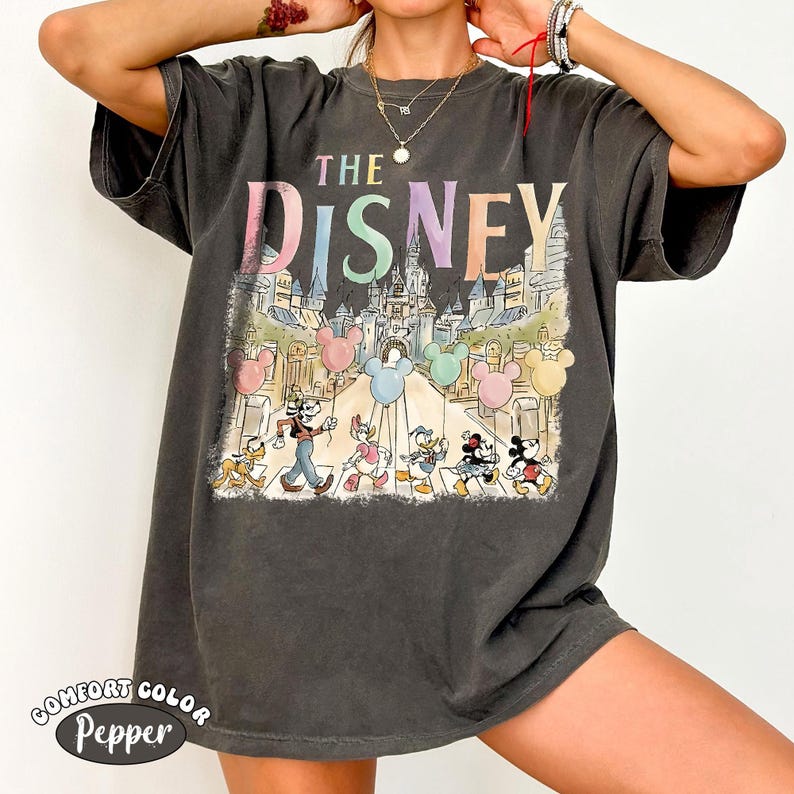 Pu&ograve; includere: T-shirt grigio scuro con grafica colorata. Il testo "THE DISNEY" &egrave; sopra un'illustrazione di personaggi Disney e un castello. La maglietta ha un aspetto vintage e sbiadito, con le parole "COMFORT COLOR Pepper" stampate in basso.