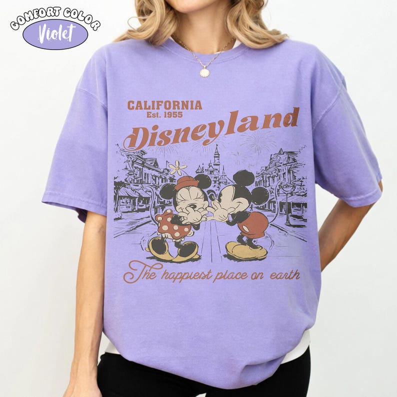 May include: A violet short-sleeve t-shirt featuring a graphic print of Mickey and Minnie Mouse in front of Disneyland. The text reads California Est. 1955, Disneyland, and The happiest place on earth. A Comfort Color Violet label is in the corner.