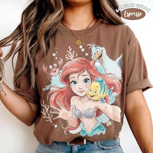 May include: Espresso-coloured t-shirt featuring a cartoon illustration of Ariel from The Little Mermaid, with Flounder, Sebastian, and Scuttle. The graphic includes coral and bubbles. The shirt is a Comfort Colors brand.