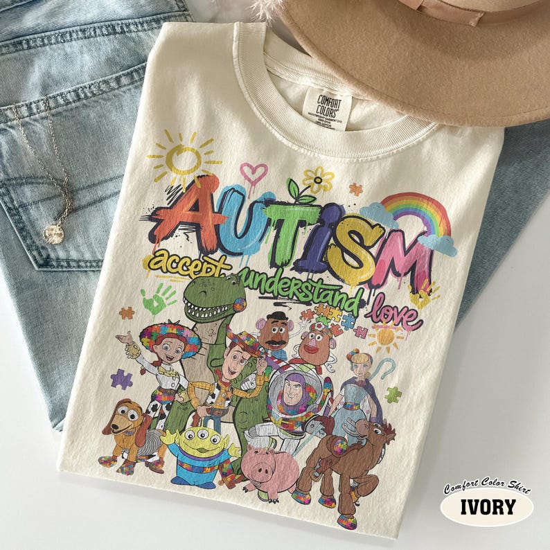 May include: An ivory t-shirt featuring a colourful graphic with the text "Autism accept understand love" and illustrations of various animated toy characters including a cowboy, space ranger, dinosaur, and potato-shaped figures with a rainbow.
