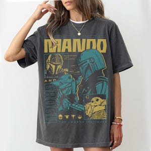 K&ouml;nnte beinhalten: Dunkelgraues T-Shirt mit einer Grafik von The Mandalorian und Grogu. Das Design enth&auml;lt den Text "MANDO" und "GROGU" sowie den Satz "WHEREVER I GO, HE GOES". Das T-Shirt hat einen Vintage-Look.