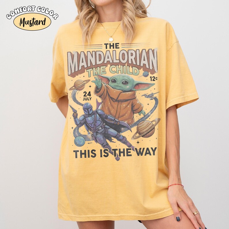May include: A mustard yellow t-shirt featuring a graphic print of The Mandalorian and The Child in space. The design includes planets, stars, and the text "The Mandalorian The Child", "24 July", "12c", and "This Is The Way". A "Comfort Color Mustard" label is visible at the top left.