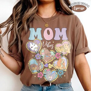 Pu&ograve; includere: T-shirt marrone con la scritta "MOM" in lettere pastello e la parola "Vibes" in corsivo. La maglietta presenta disegni a forma di cuore con personaggi dei cartoni animati e accenti floreali. La maglietta &egrave; etichettata "Comfort Color Espresso".
