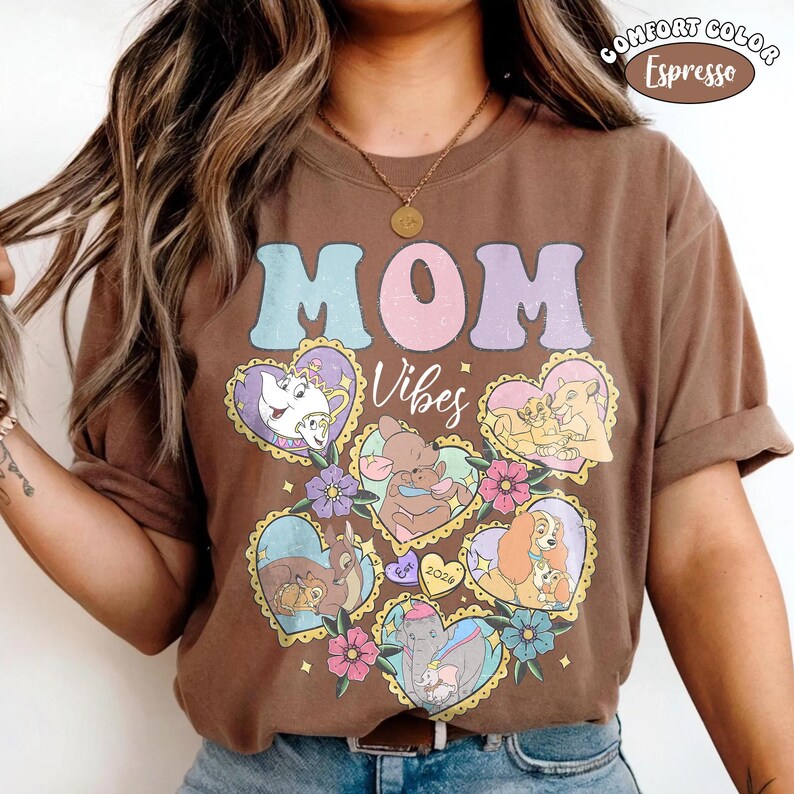 Pu&ograve; includere: T-shirt marrone con la scritta "MOM" in lettere pastello e la parola "Vibes" in corsivo. La maglietta presenta disegni a forma di cuore con personaggi dei cartoni animati e accenti floreali. La maglietta &egrave; etichettata "Comfort Color Espresso".