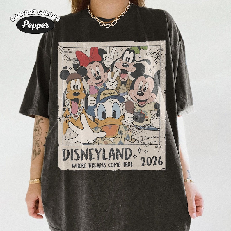 Puede incluir: Una camiseta gris carb&oacute;n con un estampado gr&aacute;fico de personajes de Disney que incluye a Mickey Mouse, Minnie Mouse, el Pato Donald, Goofy y Pluto. El texto dice Disneyland Where Dreams Come True 2026, con firmas de personajes y una etiqueta Comfort Colors Pepper.