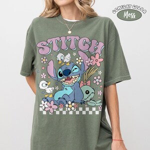 Puede incluir: Camiseta verde musgo con un gr&aacute;fico de Stitch de Lilo & Stitch, rodeado de flores y patos. La palabra "STITCH" est&aacute; en letras grandes. La camiseta est&aacute; etiquetada como "Comfort Color Moss".