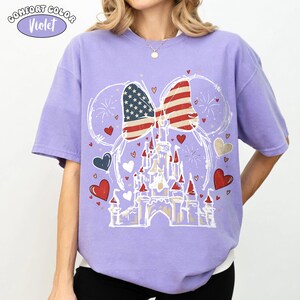 May include: A Comfort Colors violet t-shirt featuring a Minnie Mouse bow with an American flag design. The design includes a castle, hearts, and fireworks. The shirt has the text "Comfort Color Violet" in a circle.