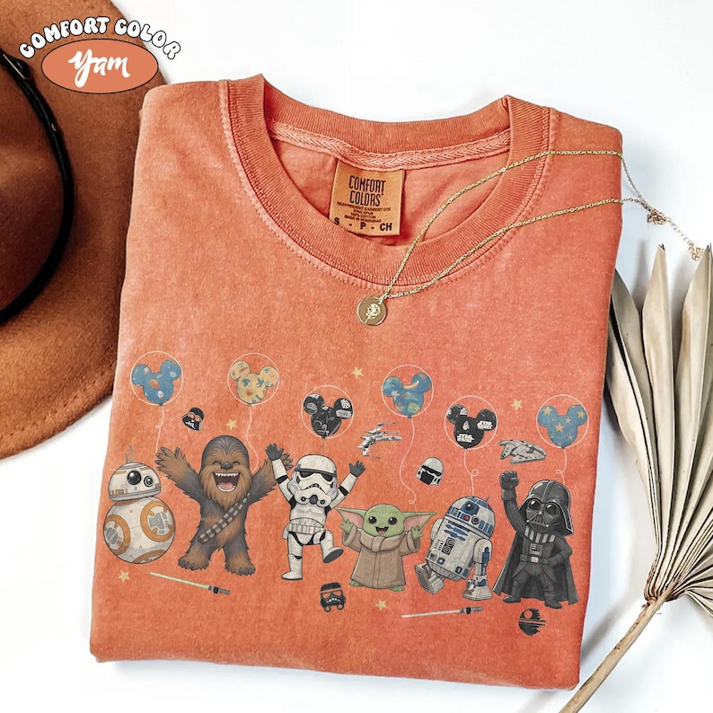 May include: Orange Comfort Colors t-shirt featuring Star Wars characters and Mickey Mouse balloons. The design includes BB-8, Chewbacca, a Stormtrooper, Grogu, R2-D2, and Darth Vader. A gold necklace and a brown hat are also visible.