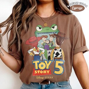 Pu&ograve; includere: T-shirt color espresso con una grafica dei personaggi del film d'animazione Toy Story. Il design include Woody, Buzz Lightyear, Jessie e altri personaggi, con la scritta "TOY STORY 5" e il logo Disney Pixar.