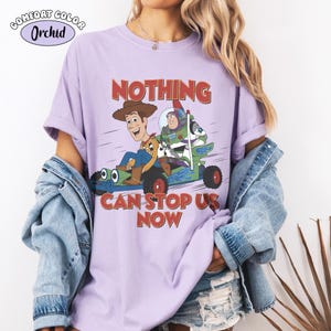May include: A lilac Comfort Colors t-shirt featuring a cartoon image of Woody and Buzz Lightyear in a go-kart. The text "NOTHING CAN STOP US NOW" is printed in red. The shirt has short sleeves and a crew neck.