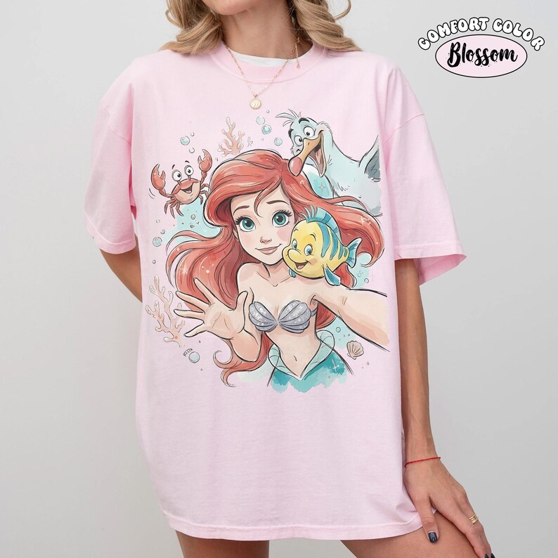 May include: Pale pink Comfort Colors t-shirt with a cartoon illustration of Ariel from The Little Mermaid, with Flounder, Sebastian, and Scuttle. The graphic shows Ariel taking a selfie. The text "Comfort Color Blossom" is printed on the upper right.