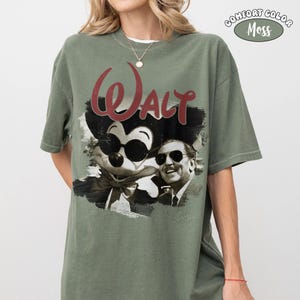 Puede incluir: Camiseta verde oliva Comfort Colors con un gr&aacute;fico de Mickey Mouse con gafas de sol y Walt Disney. La palabra "Walt" est&aacute; en escritura roja encima de la imagen. La camiseta tiene mangas cortas y cuello redondo.