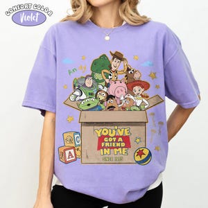 K&ouml;nnte beinhalten: Ein violettes Comfort Colors T-Shirt mit einer Grafik von Toy Story-Charakteren. Das Design beinhaltet eine Box mit dem Text "YOU'VE GOT A FRIEND IN ME SINCE 1995" und Spielzeugbl&ouml;cken.