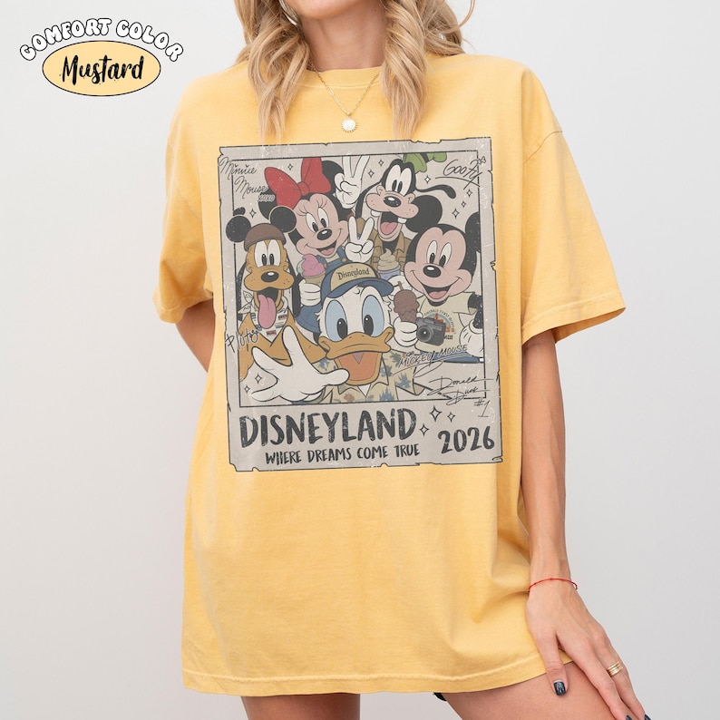 Puede incluir: Una camiseta de manga corta color amarillo mostaza con un estampado gr&aacute;fico rectangular de personajes de Disney, incluidos Mickey Mouse, Minnie Mouse, Donald Duck, Goofy y Pluto. El texto dice Disneyland Where Dreams Come True 2026.