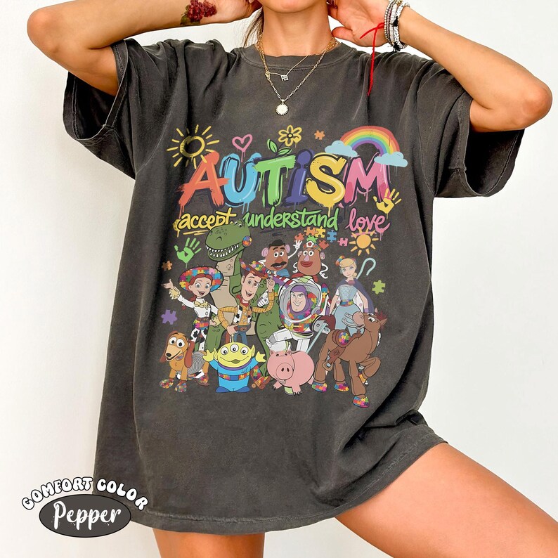 May include: A charcoal grey short-sleeve t-shirt featuring a colourful graphic print with the text "AUTISM" and "accept understand love". The design includes illustrations of various animated toy characters, a rainbow, a sun, and puzzle pieces.