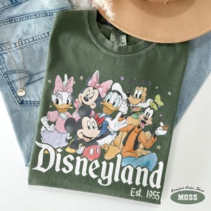 Pu&ograve; includere: Una t-shirt girocollo verde oliva con una stampa grafica di Topolino, Minni, Paperino, Paperina e Pippo. Il testo "Disneyland" e "Est. 1955" &egrave; stampato in bianco sotto i personaggi, circondato da piccole stelle.