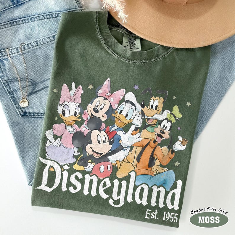 Pu&ograve; includere: Una t-shirt girocollo verde oliva con una stampa grafica di Topolino, Minni, Paperino, Paperina e Pippo. Il testo "Disneyland" e "Est. 1955" &egrave; stampato in bianco sotto i personaggi, circondato da piccole stelle.