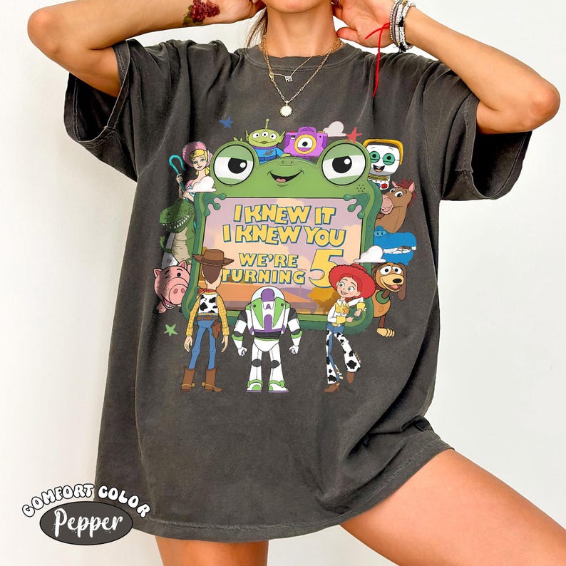 Puede incluir: Una camiseta de manga corta gris carb&oacute;n con un estampado gr&aacute;fico de varios personajes animados rodeando un marco verde con forma de rana. El texto dentro del marco dice I KNEW IT I KNEW YOU WE'RE TURNING 5. La camiseta tiene cuello redondo.