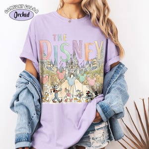 Pu&ograve; includere: Una maglietta lilla Comfort Colors con una grafica colorata della parola "DISNEY" sopra un'illustrazione di un castello. Topolino e i suoi amici sono raffigurati mentre attraversano un passaggio pedonale. La maglietta ha le maniche arrotolate.