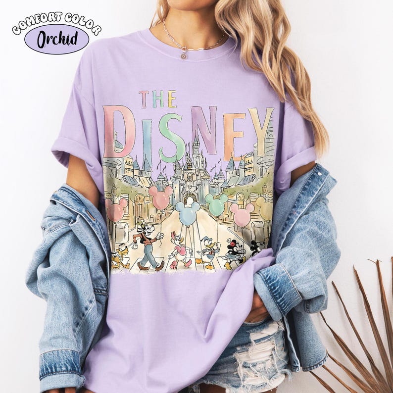 Pu&ograve; includere: Una maglietta lilla Comfort Colors con una grafica colorata della parola "DISNEY" sopra un'illustrazione di un castello. Topolino e i suoi amici sono raffigurati mentre attraversano un passaggio pedonale. La maglietta ha le maniche arrotolate.